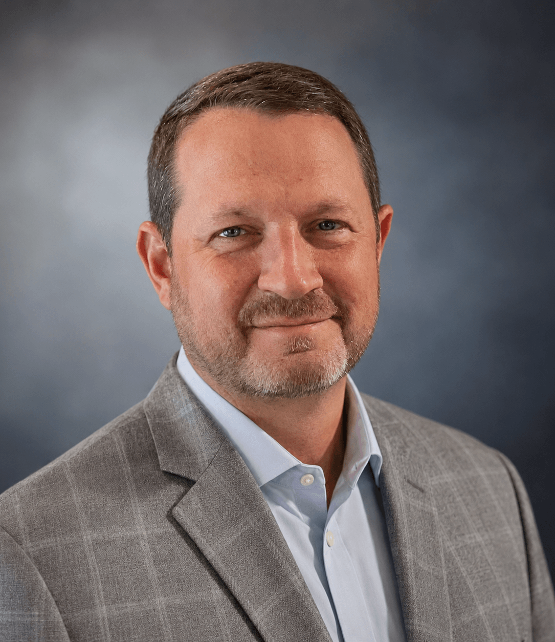 A middle-aged man with short brown hair and a beard smiles directly at the camera. He is wearing a light blue collared shirt and a grey plaid blazer.